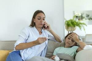 madre misurazione temperatura di sua malato ragazzo. malato bambino con alto febbre posa nel letto e madre Tenere termometro. madre con cellula Telefono chiamata per medico foto