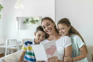 gioioso giovane madre e poco bambini seduta su divano e Abbracciare una persona mentre lettura auguri e Congratulazioni con vacanza nel presentata cartolina a casa foto