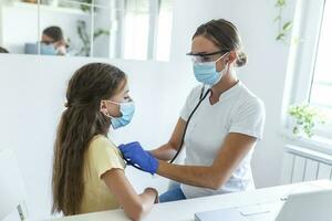 adorabile ragazza è seduta a sua medico ufficio. sua medico è utilizzando un' stetoscopio per ascolta per sua il petto. tutti e due sua e sua medico siamo indossare un' viso maschera per impedire il diffusione di germi. foto