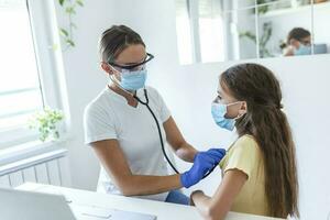 adorabile ragazza è seduta a sua medico ufficio. sua medico è utilizzando un' stetoscopio per ascolta per sua il petto. tutti e due sua e sua medico siamo indossare un' viso maschera per impedire il diffusione di germi. foto