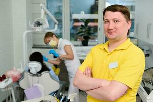 fiducioso contento maschio dentista nel giallo uniforme in piedi nel dentale ufficio con braccia piegato, sorridente guardare a telecamera foto
