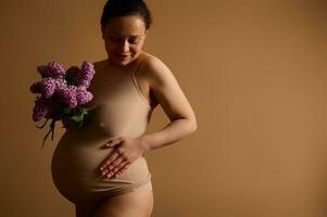 attraente incinta donna sorridente, toccante sua gonfiarsi, in posa con un' mazzo di fioritura lillà, vestito nel beige biancheria intima foto