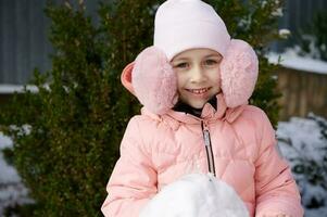 caucasico poco bambino ragazza nel caldo inverno Abiti e rosa soffice paraorecchie, sorrisi a telecamera, edificio pupazzo di neve all'aperto foto