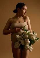 gravido donna nel beige biancheria intima, Tenere mano su sua incinta gonfiarsi, guardare a parte, in posa con mazzo di fioritura lillà foto