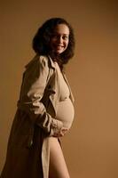 attraente elegante gravido donna nel biancheria intima sotto beige trincea cappotto, Tenere sua incinta gonfiarsi, sorridente guardare a parte. foto