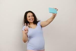 bellissimo incinta donna Spettacoli pace cartello, prende autoscatto su smartphone, condivisione contento gravidanza stile di vita su sociale media foto