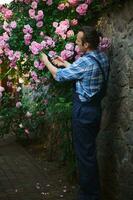 pieno lunghezza ritratto maschio giardiniere floricoltore nel blu giardinaggio uniforme, tendente impianti nel il cortile di dimora foto