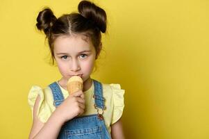 avvicinamento poco bambino ragazza leccata notizia in anticipo di ghiaccio crema nel cialda cono, guardare a telecamera, isolato al di sopra di giallo sfondo foto