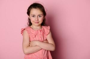 contento bambino ragazza indossare rosa vestire, in piedi con braccia piegato al di sopra di rosa isolato sfondo, sorridente guardare a telecamera foto