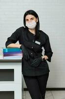 femmina medico nel nero medico uniforme, in piedi con un' pila di medico letteratura al di sopra di bianca mattone parete sfondo foto