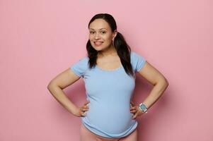 bellissimo incinta donna nel blu maglietta, con grande gonfiarsi, sorridente guardare a telecamera, isolato rosa sfondo. gravidanza foto