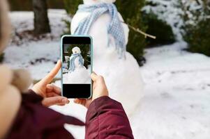 avvicinamento mani hold smartphone nel vivere Visualizza modalità, Fotografare pupazzo di neve con blu scialle nel neve coperto natura sfondo foto