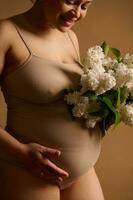 avvicinamento contento incinta donna madre nel biancheria intima con mazzo di bianca lilla fiori, tocchi e colpi sua grande pancia foto