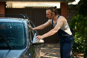 attraente muscolare ispanico uomo nel blu tuta, manualmente lavaggio il suo auto con spugna e schiumoso detergente nel Giardino dietro la casa foto