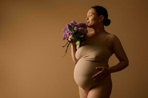 gravidanza. maternità. gravidanza. corpo positività. gravido donna nel biancheria intima, con mazzo di lillà su beige sfondo foto