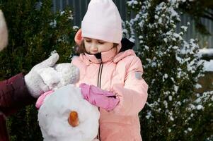 carino poco bambino ragazza nel caldo rosa giù giacca, godendo fabbricazione un' pupazzo di neve nel un' Giardino dietro la casa, la spesa contento inverno vacanze foto