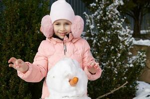 bello poco ragazzo ragazza indossare caldo rosa giù giacca e soffice paraorecchie, sorrisi a telecamera, edificio pupazzo di neve nel Giardino dietro la casa foto