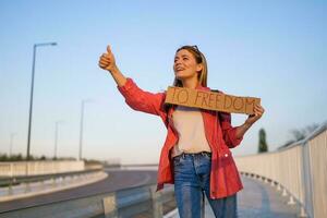 donna è autostop su ciglio della strada provando per fermare macchina. lei è Tenere cartone con iscrizione. foto