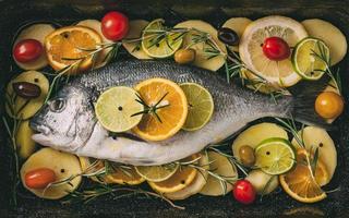 orata pesce in teglia pronto per essere infornato con patate, rosmarino, limone, arancia, olive, pomodori, cipolla e lime. orata fresca, preparazione di pesce dorade