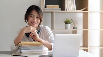 donna asiatica sorridente che lavora alla scrivania con libri e computer portatile foto