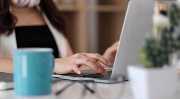 mani della donna d'affari che digitano, navigando in Internet sulla tastiera del computer portatile con smartphone mobile, auricolari, una tazza di caffè sul tavolo dell'ufficio in legno presso l'home office. studentessa che impara online foto