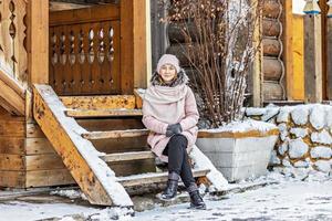 giovane donna vestita calorosamente in posa sotto il portico di una casa in legno nel villaggio. vacanze invernali in campagna