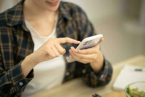 contento bellissimo asiatico donna con Telefono mangiare fresco insalata a tavolo nel cucina e utilizzando applicazione sms su smartphone mentre mangiare salutare cibo foto