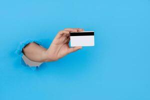 mano braccio attraverso carta e Tenere credito carta per shopping isolato al di sopra di blu sfondo. foto
