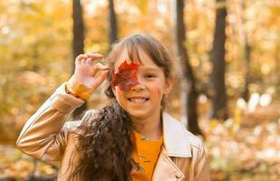 vicino ritratto di un' poco ragazza. lei coperture sua occhio con un' acero autunno foglia. autunno stagione e bambini concetto. foto