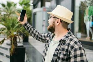 barbuto uomo prende autoscatto attraverso smartphone passeggiate nel centro pose contro moderno città edificio gode tempo libero volta. persone urbano stile di vita e tecnologia concetto foto