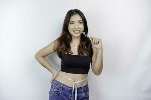 ritratto di contento giovane asiatico donna indossare corpo misurazione nastro in giro vita Tenere cucchiaio e forchetta isolato al di sopra di bianca sfondo foto