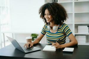 bellissimo africano donna utilizzando il computer portatile e tavoletta mentre seduta a sua Lavorando posto. concentrato a opera nel casa ufficio foto