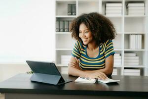 bellissimo africano donna utilizzando il computer portatile e tavoletta mentre seduta a sua Lavorando posto. concentrato a opera nel casa ufficio foto