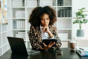 giovane africano donna digitando su tavoletta e il computer portatile mentre seduta a il Lavorando nero tavolo ufficio foto