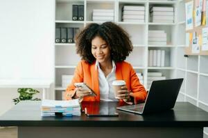 giovane africano donna digitando su tavoletta e il computer portatile mentre seduta a il Lavorando nero tavolo ufficio foto