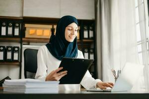 giovane Arabo femmina imprenditore indossare un' hijab Lavorando in linea con un' il computer portatile a ufficio foto