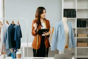 moda progettista donna parlando inteligente Telefono e utilizzando il computer portatile con digitale tavoletta computer nel moderno studio il Abiti sospeso su il cremagliere mattina leggero foto