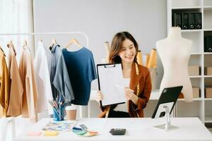 asiatico sarto donna Lavorando su Abiti nel sartoria atelier. bellissimo giovane femmina moda progettista Sorridi e dopo successo nel studio foto