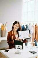 asiatico sarto donna Lavorando su Abiti nel sartoria atelier. bellissimo giovane femmina moda progettista Sorridi e dopo successo nel studio foto