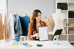 asiatico sarto donna Lavorando su Abiti nel sartoria atelier. bellissimo giovane femmina moda progettista Sorridi e dopo successo nel studio foto