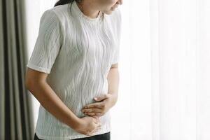 giovane asiatico donna stomaco dolore seduta su letto a casa, Salute problema infiammazione nel corpo, periodo ciclo giorno di mensile, mestruazione concetto. foto