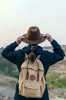 escursionista donna a partire dal indietro con blu giacca e zaino, detiene sua fricchettone cappello, mentre guardare a il orizzonte. escursione e viaggio concetto. foto