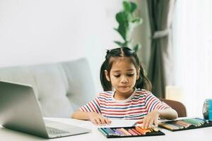 carino poco asiatico ragazza utilizzando il computer portatile e fare compiti a casa a casa. foto