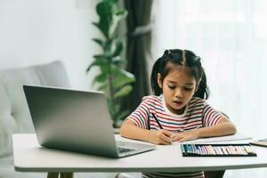 asiatico poco ragazza utilizzando il computer portatile e fare compiti a casa nel vivente camera a casa foto