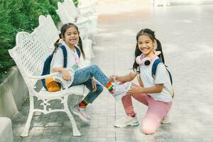 contento asiatico poco ragazze con zaino e libri seduta su il panchina foto