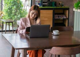 donna d'affari indossare un' completo da uomo rosa seduta a il caffè negozio e utilizzando il computer portatile conferenza video chiamata per parlare in linea incontro e utilizzando tavoletta matita per Scrivi rapporto su contabilità finanza attività commerciale. foto