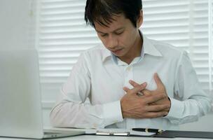 mano hold il petto con cuore attacco sintomi, asiatico uomo Lavorando difficile avere il petto dolore causato di cuore patologia, perdere, dilatazione, allargata coronario cuore, stampa su il il petto con un' doloroso espressione foto