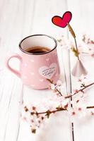caffè in tazza rosa, fiori e cuore come decorazione, su fondo di legno bianco foto