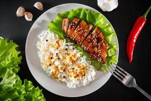 pollo teriyaki con semi di sesamo, lattuga e riso su un piatto bianco. piatto con una forchetta su sfondo nero foto