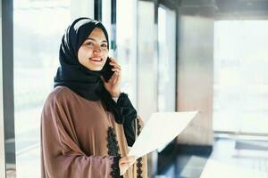 giovane musulmano donna nel hijab parlando su Telefono e Tenere carta foto